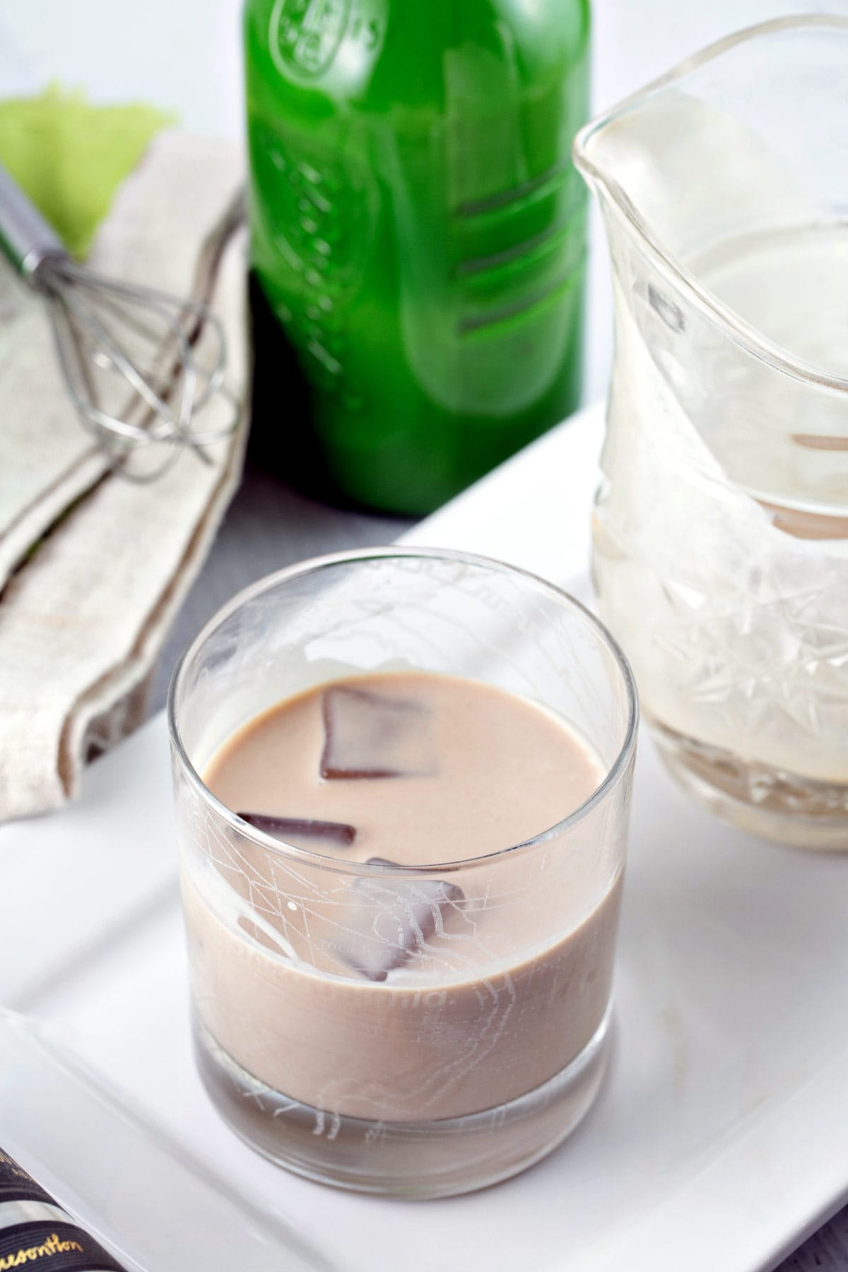 lowball glass with ice cubes and homemade irish cream in front of a green glass bottle.