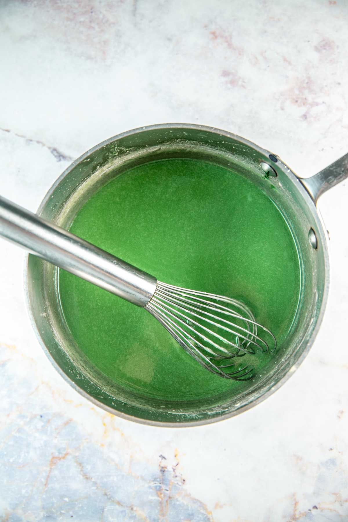 Green frosting being cooked in a silver saucepan with a silver whisk. 