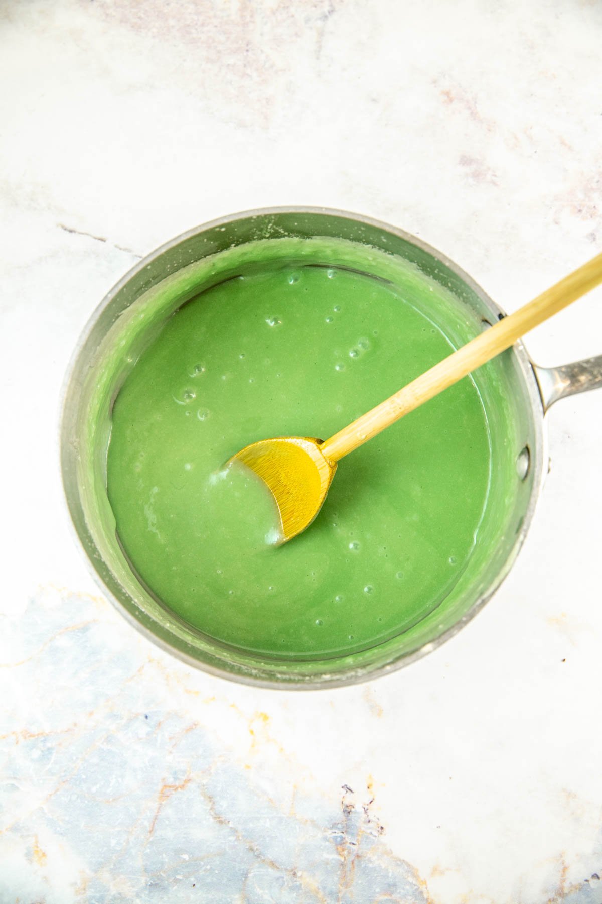 Green frosting being stirred with a wooden spoon in a saucepan. 