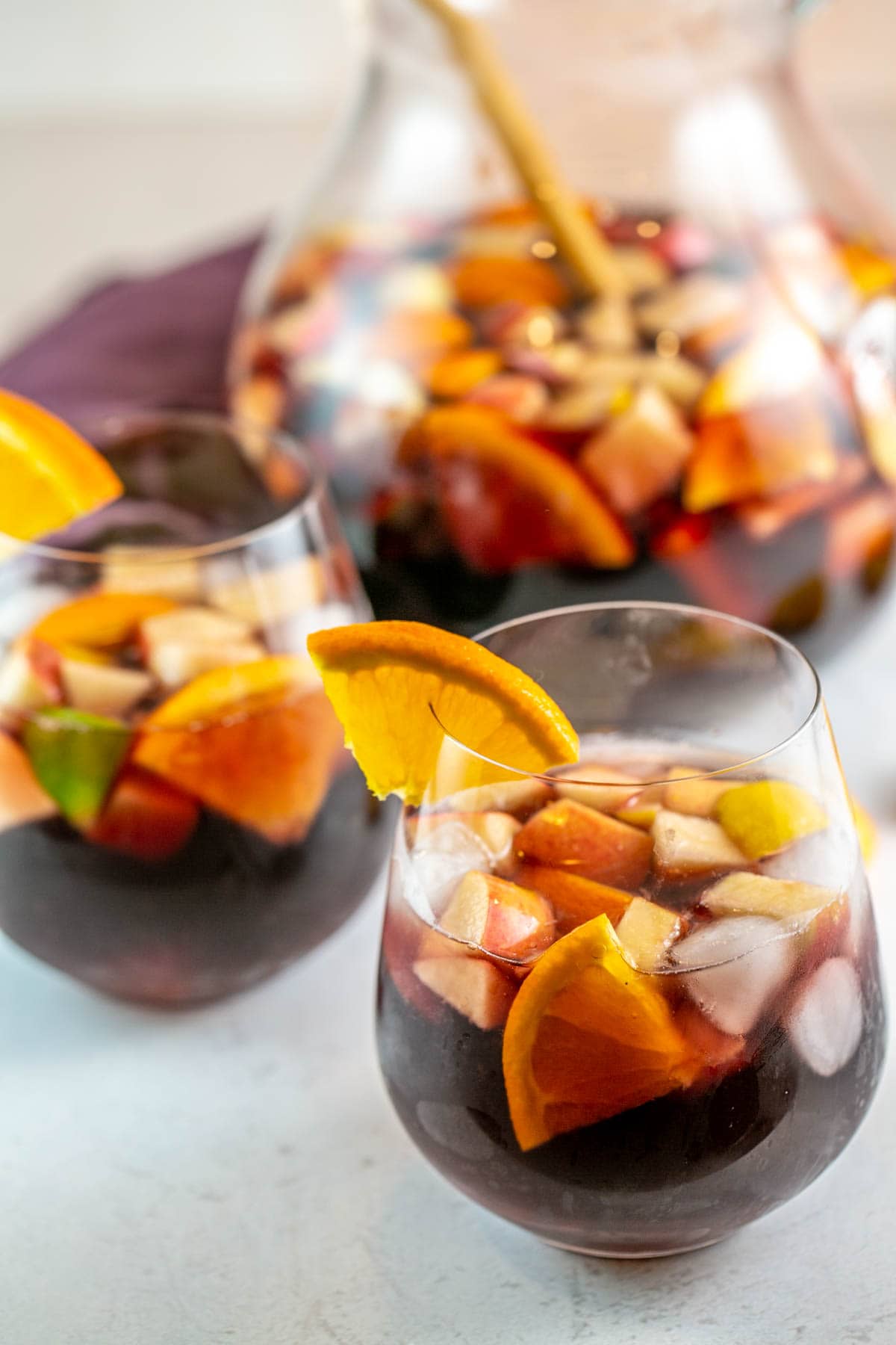 two glasses of sangria full of chopped fruit with the pitcher visible in the background