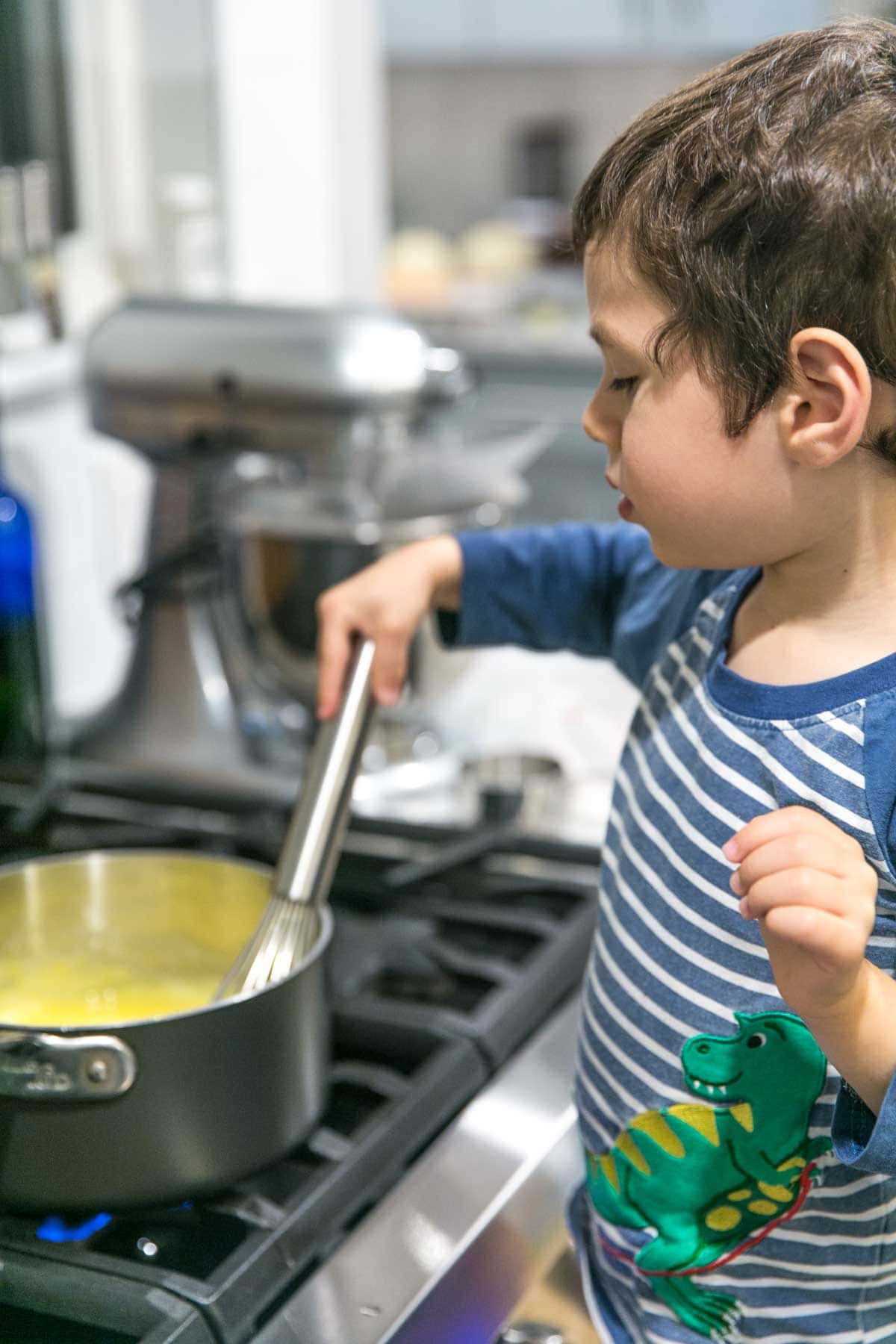 a 4 year old boy standing at the stove whisking a pot full of lemon curd