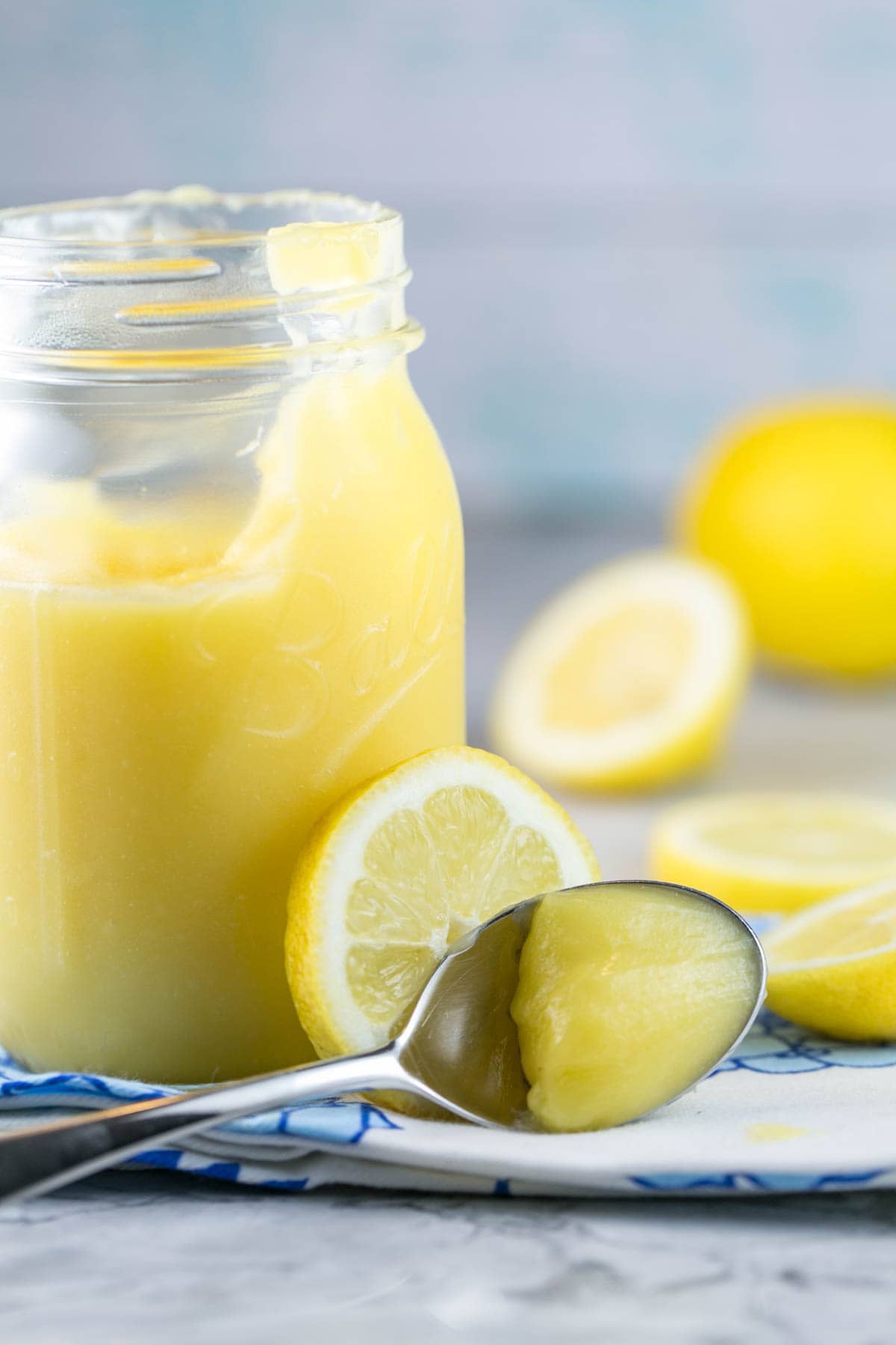 a spoon filled with lemon curd resting against a sliced lemon and a jar filled with lemon curd