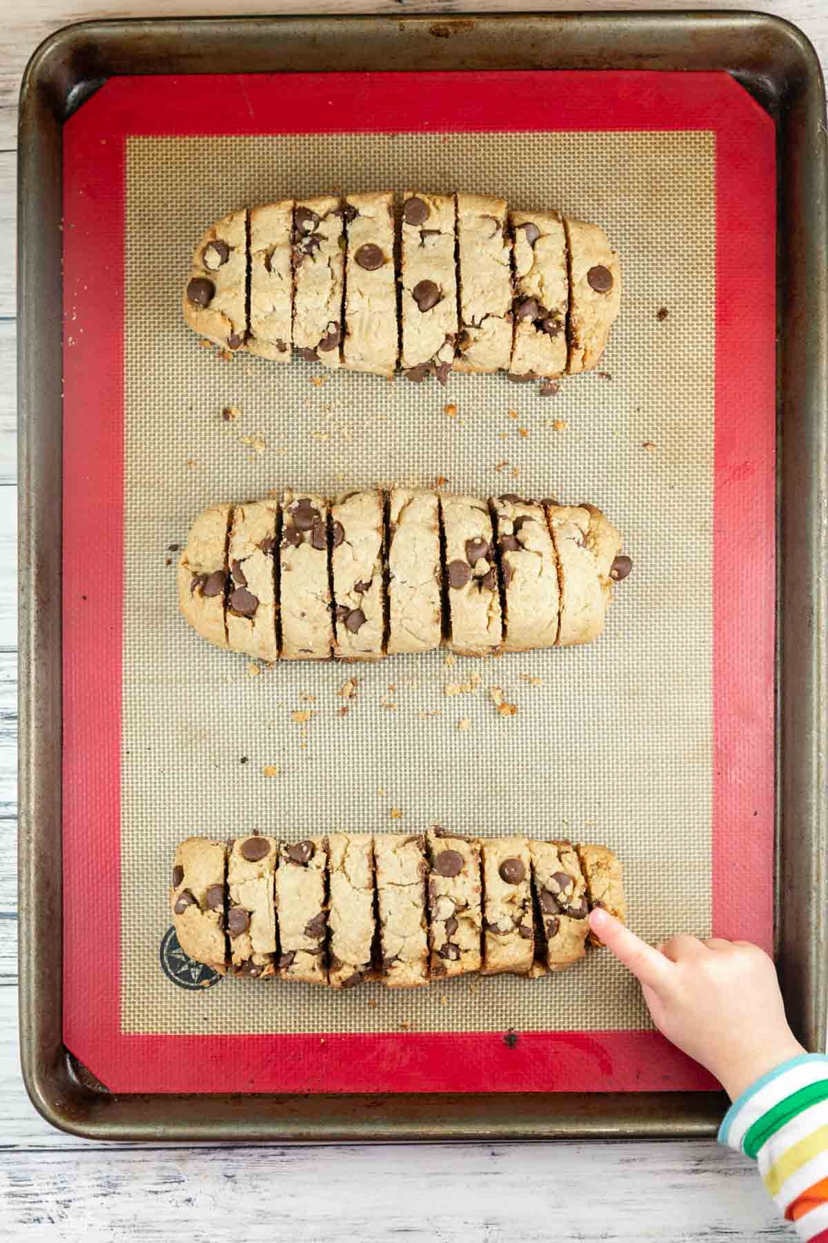 three baked loaves of mandelbrot sliced into cookies with a toddler hand reaching out to grab a cookie