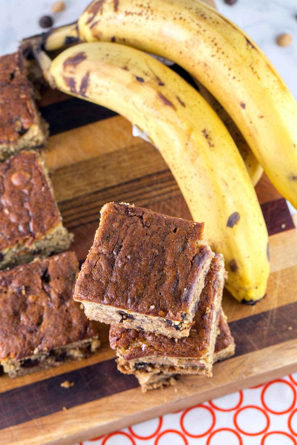 banana bars on a wooden cutting board next to a stack of ripe bananas