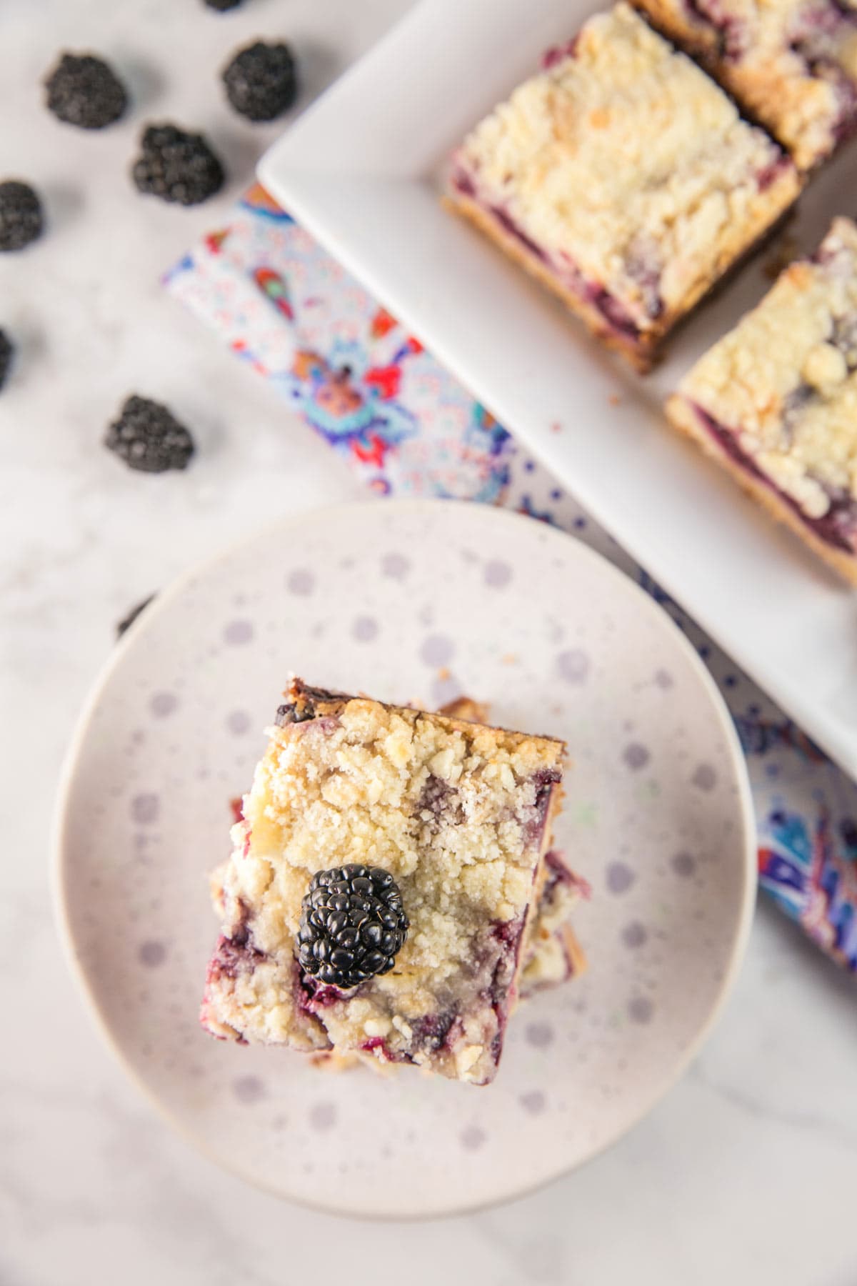 a stack of blackberry pie bars on a speckled plate with blackberries scattered in the background