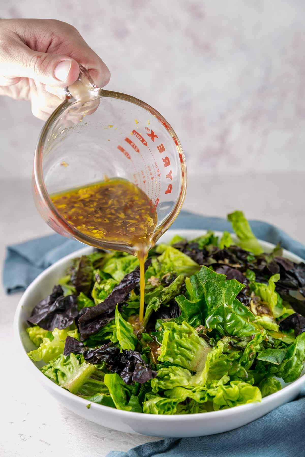 pouring a mustard shallot vinaigrette dressing from a glass measuring cup over a salad of leafy greens