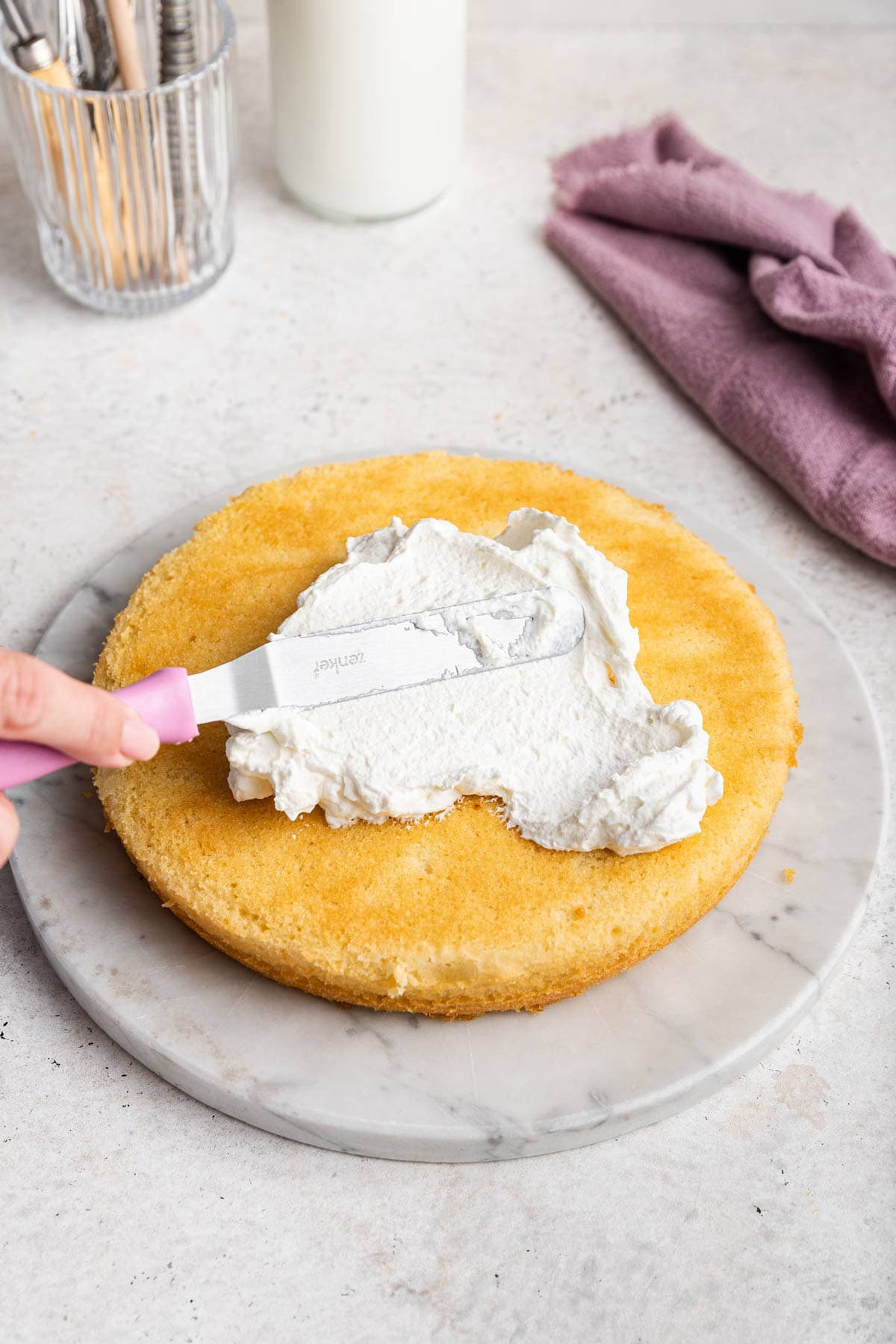 spreading whipped cream frosting on a layer of vanilla cake