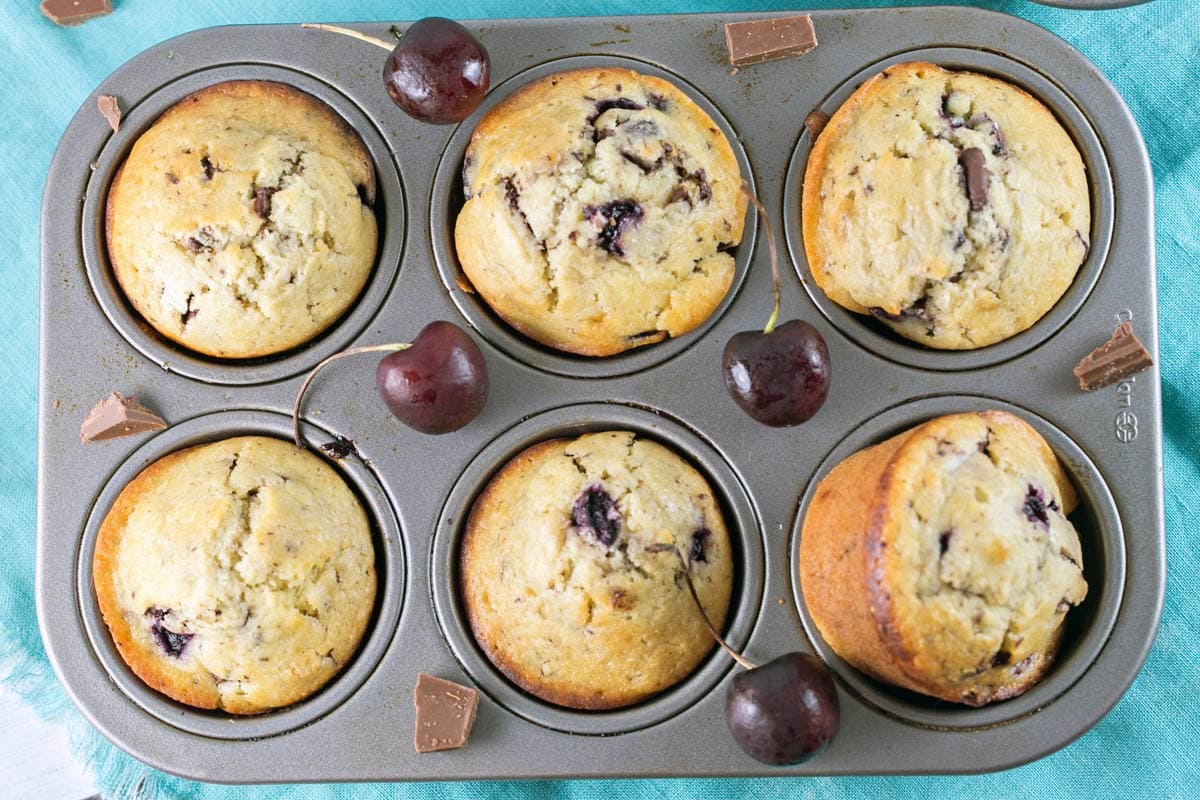 six cherry chocolate chunk muffins in a muffin tin surrounded by fresh cherries and chunks of chocolate