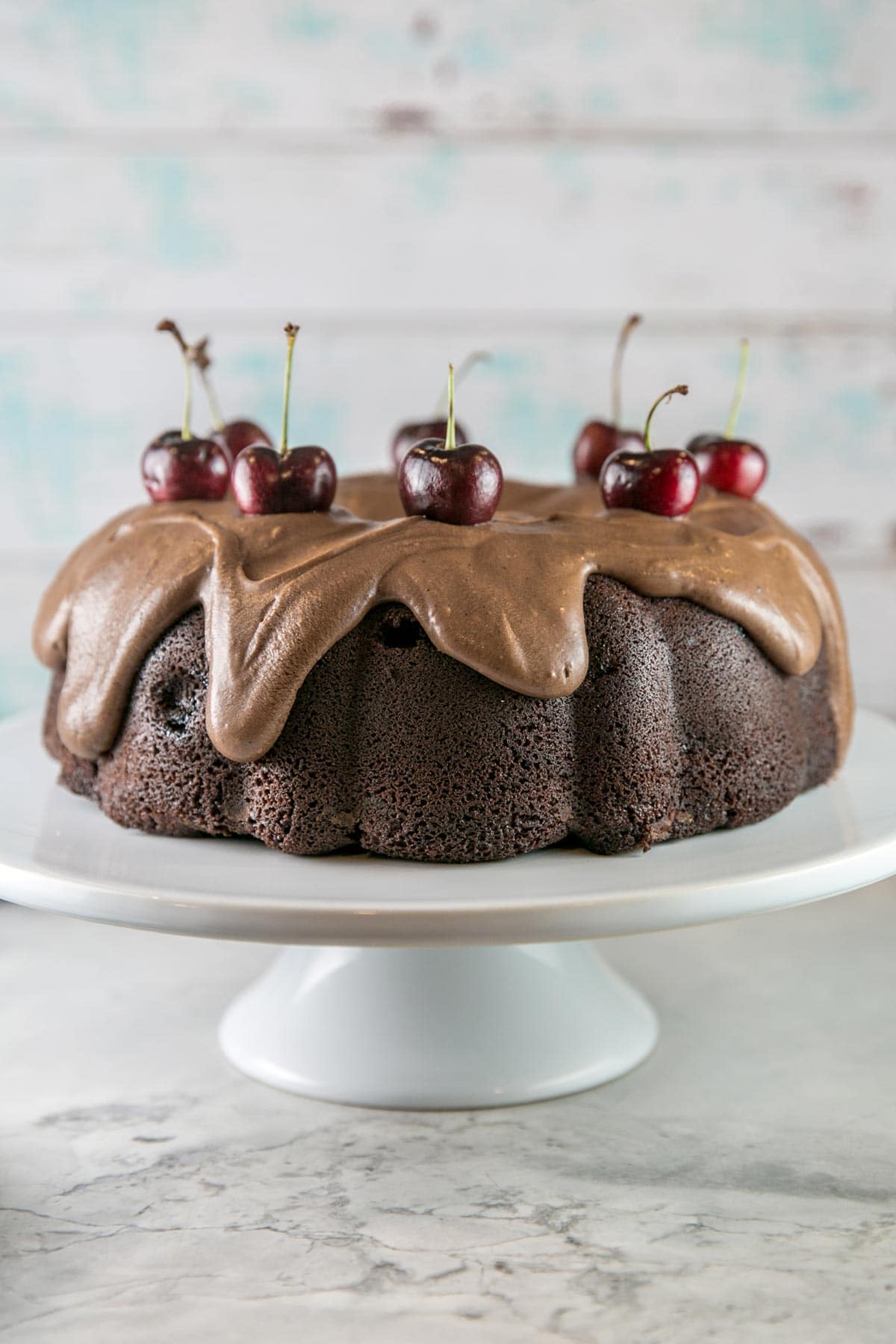 a chocolate cherry bundt cake covered in fudge frosting and topped with fresh cherries on a white cake stand