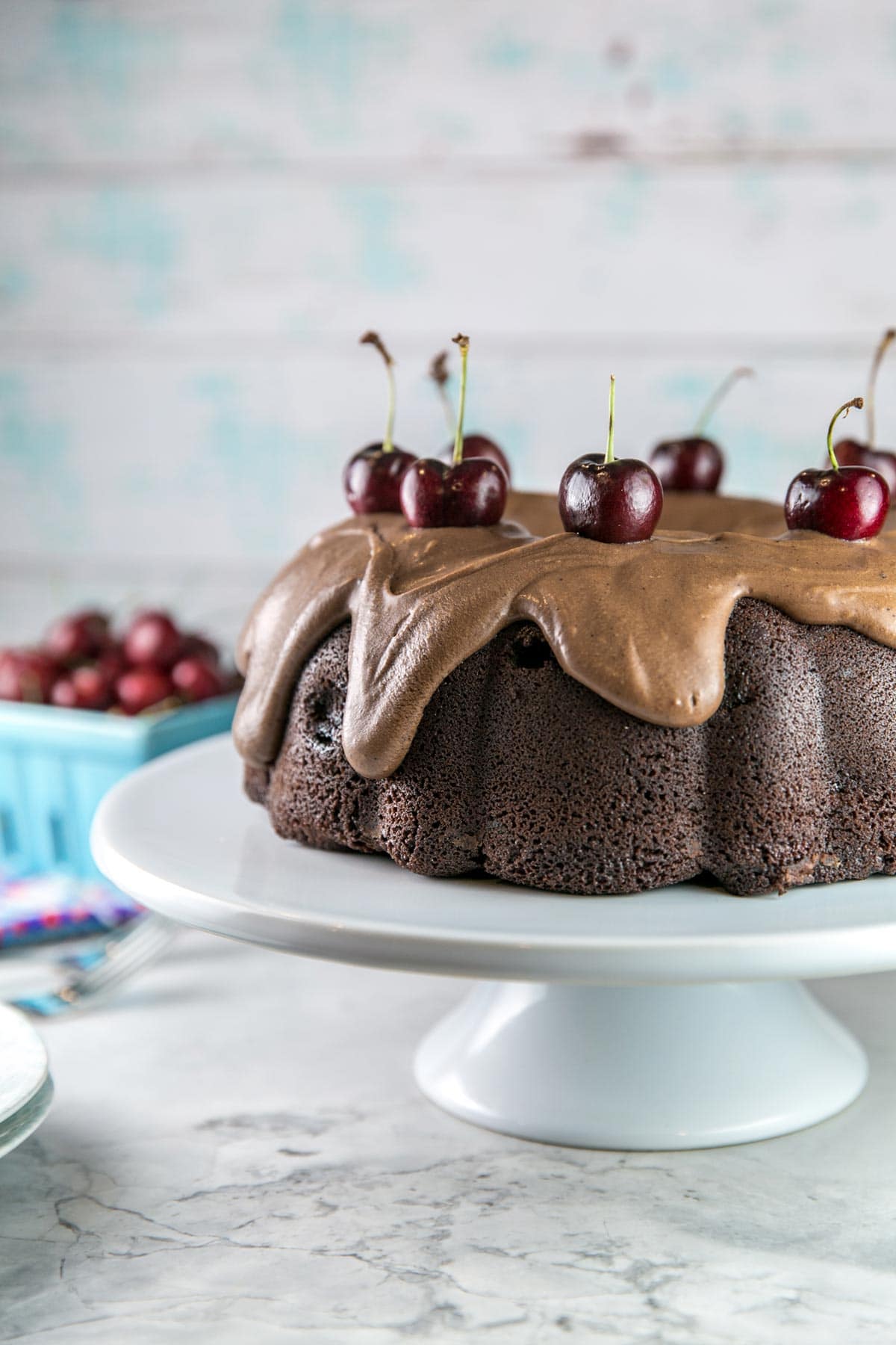 close view of an uncut chocolate bundt cake decorated with thick chocolate frosting and fresh cherries