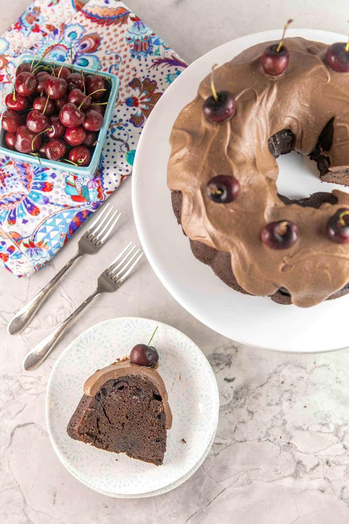 a chocolate cherry bundt cake decorated with fresh cherries next to a slice of the cake and a bowl of fresh cherries