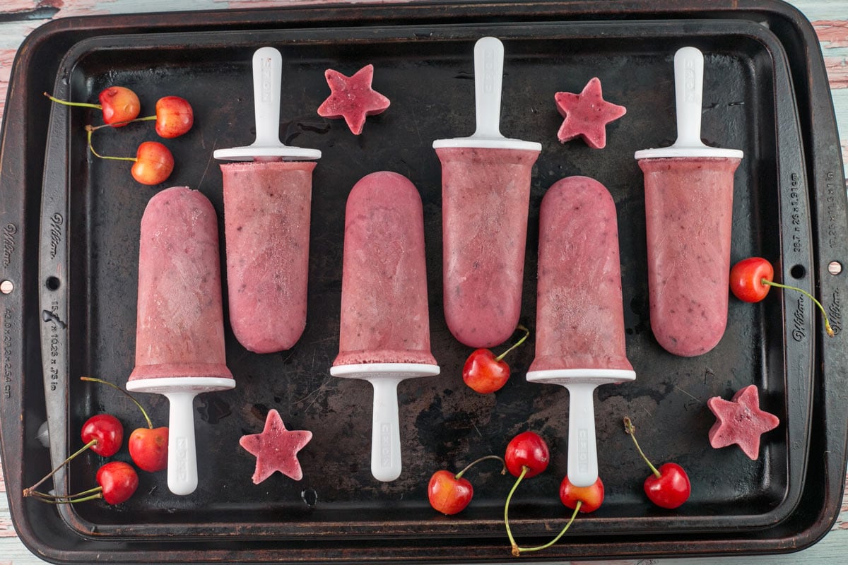 six pink-colored popsicles and fresh cherries on a metal sheet pan