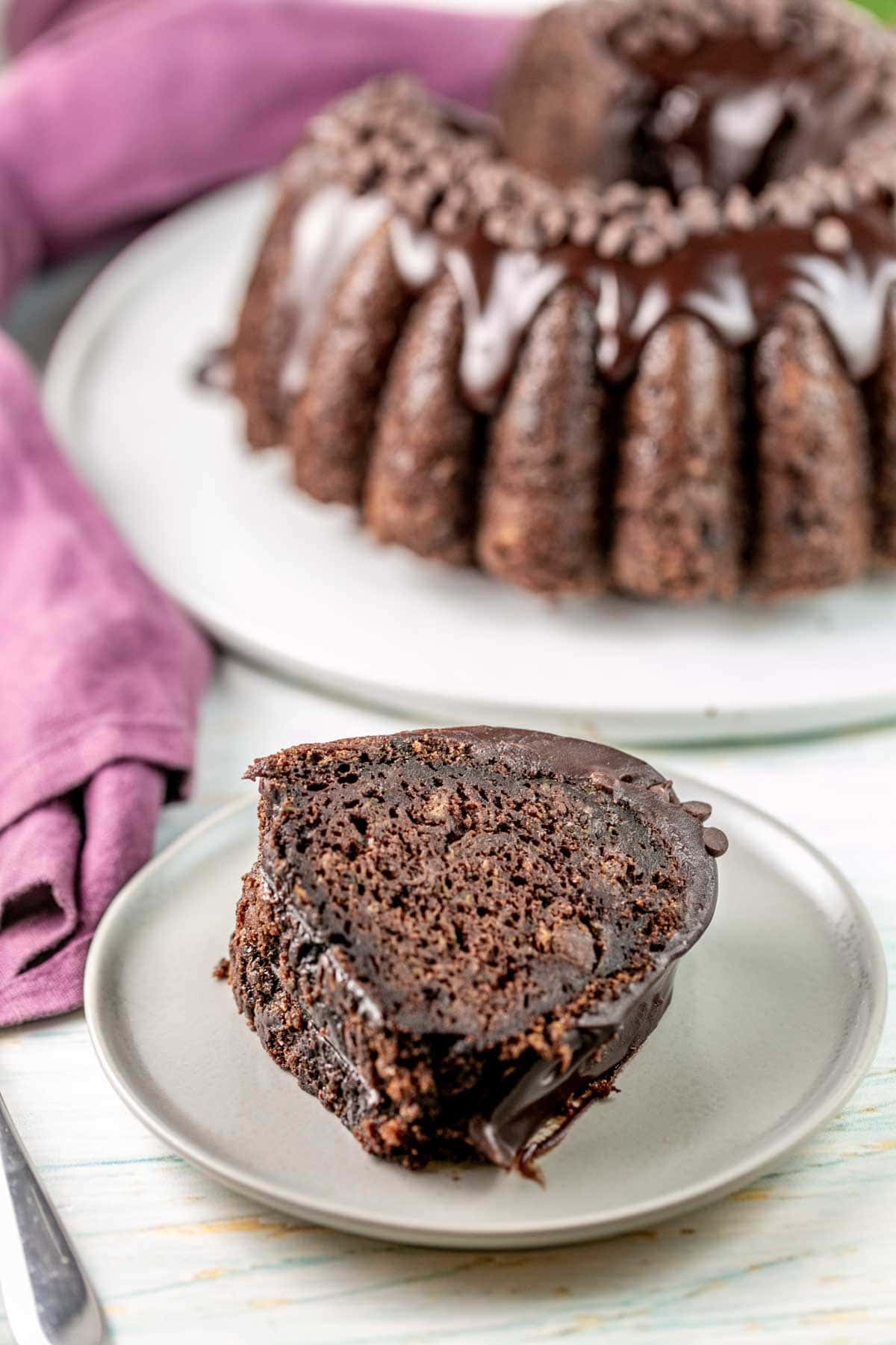 Slice of zucchini cake on a plate with whole cake on a platter in the background. 