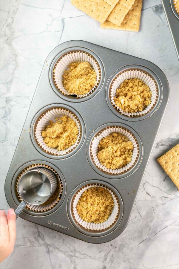 Hand pushing down the graham cracker crust in the cupcake liners. 