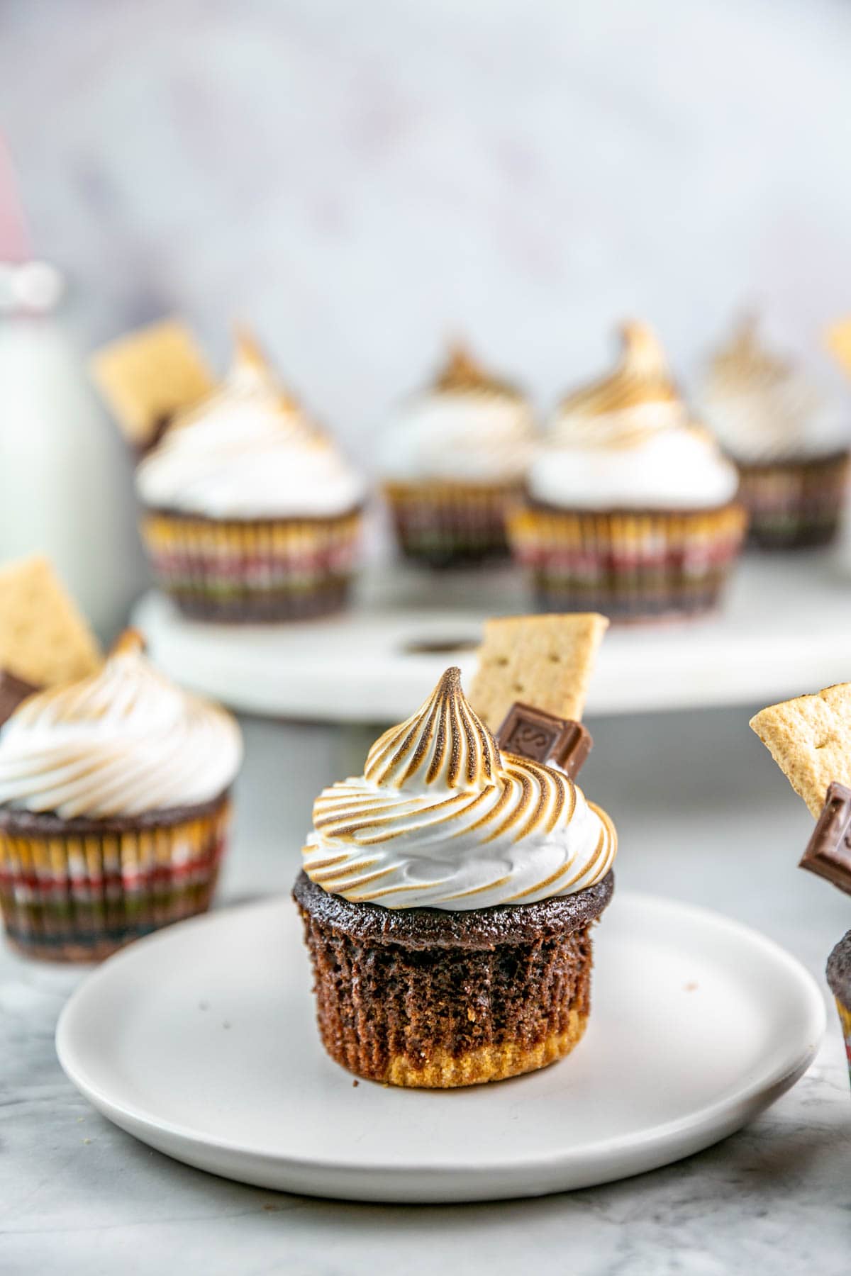 Smores cupcake on a white plate with a piece of chocolate and graham cracker on top. 
