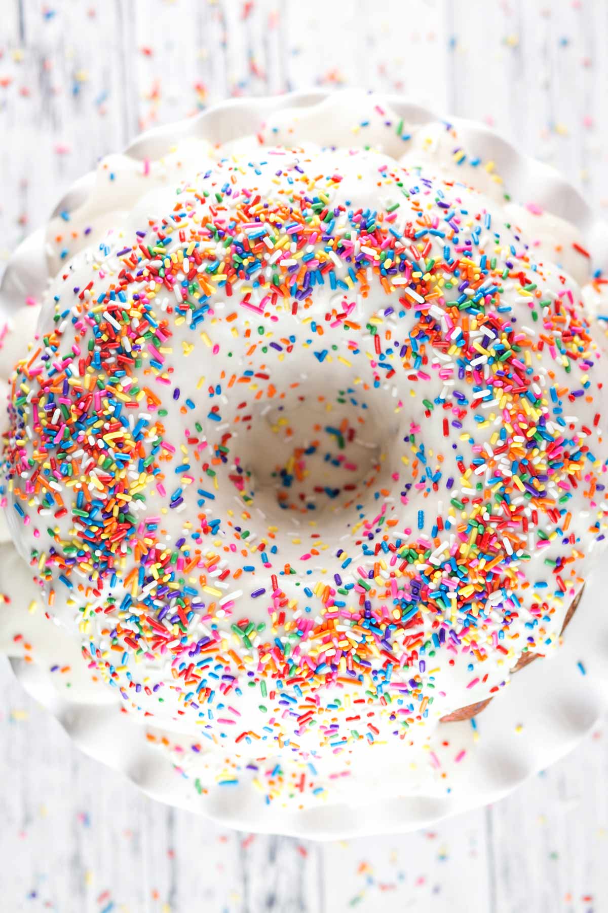 vanilla cake with glaze covered in colorful sprinkles on a white cake stand.