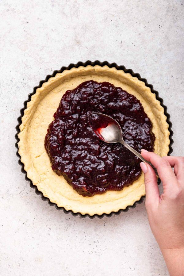 a hand holding a spoon spreading jam in a shortbread crust.