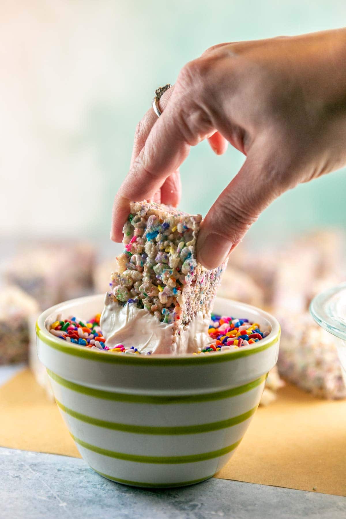 a hand dipping a rice krispie treat into a bowl full of sprinkles.