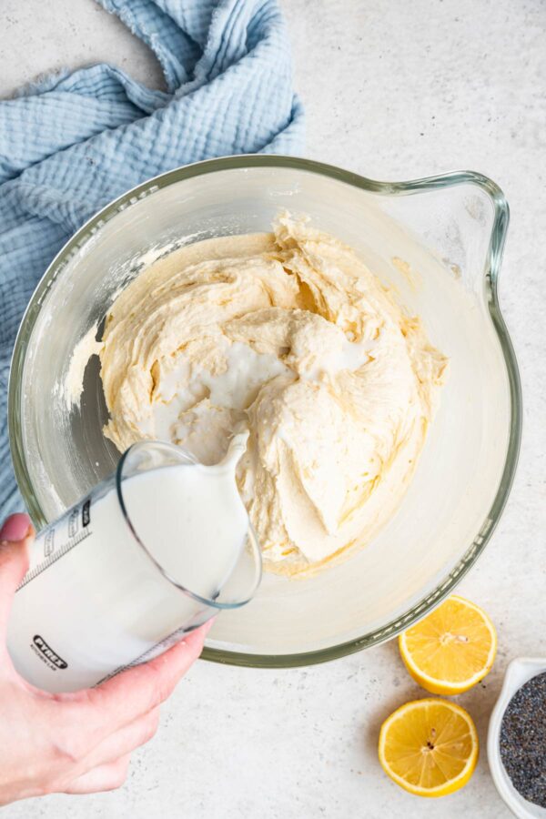 a hand pouring milk into a lemon poppy seed cake batter.