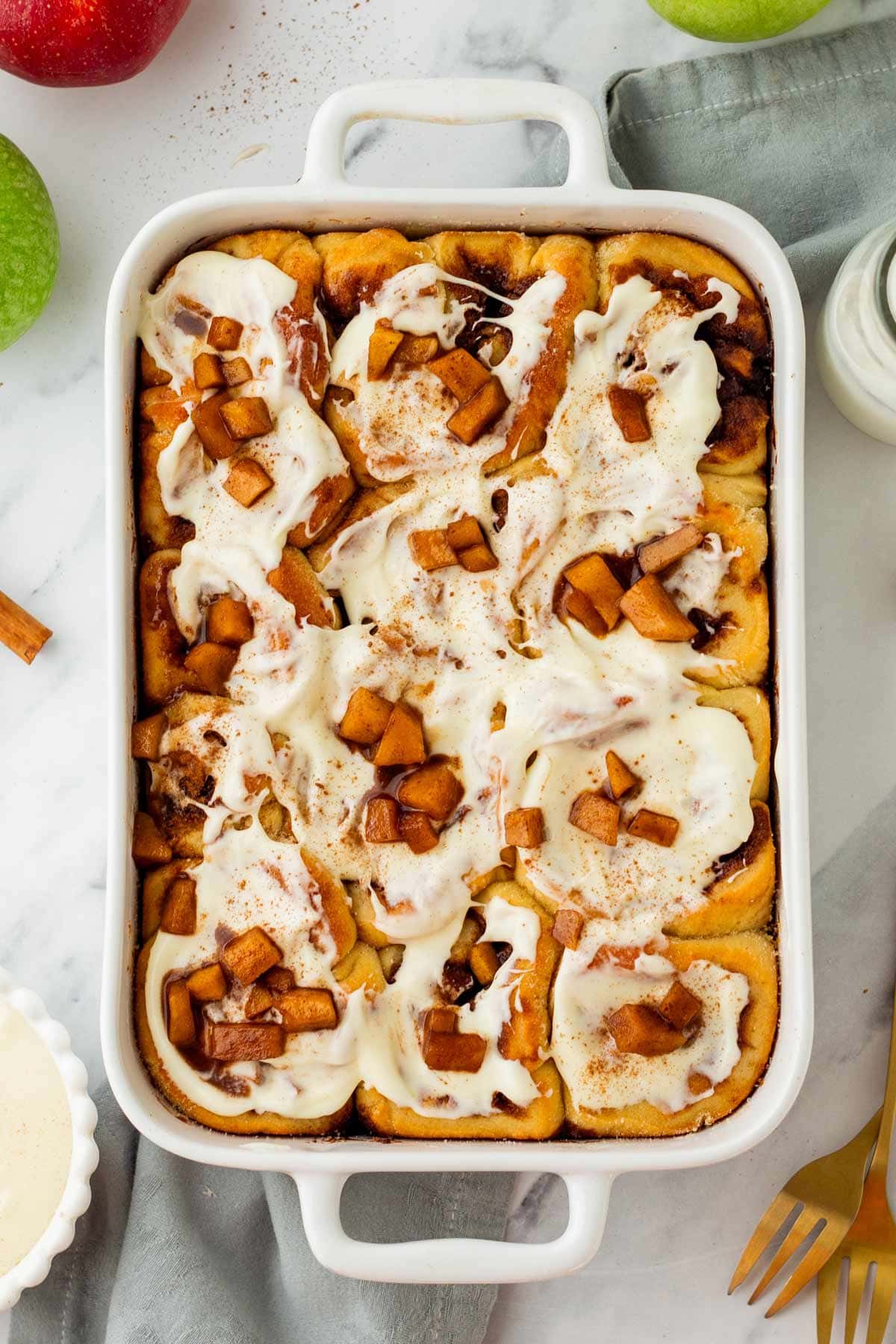 homemade apple pie cinnamon rolls covered with cream cheese frosting in a white ceramic serving dish.