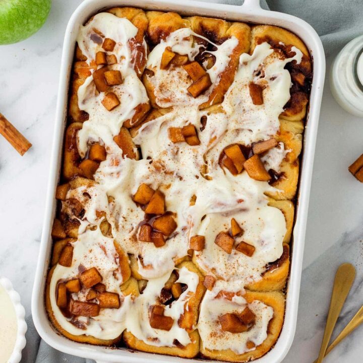 pan of freshly baked cinnamon rolls covered with frosting and spiced apples.