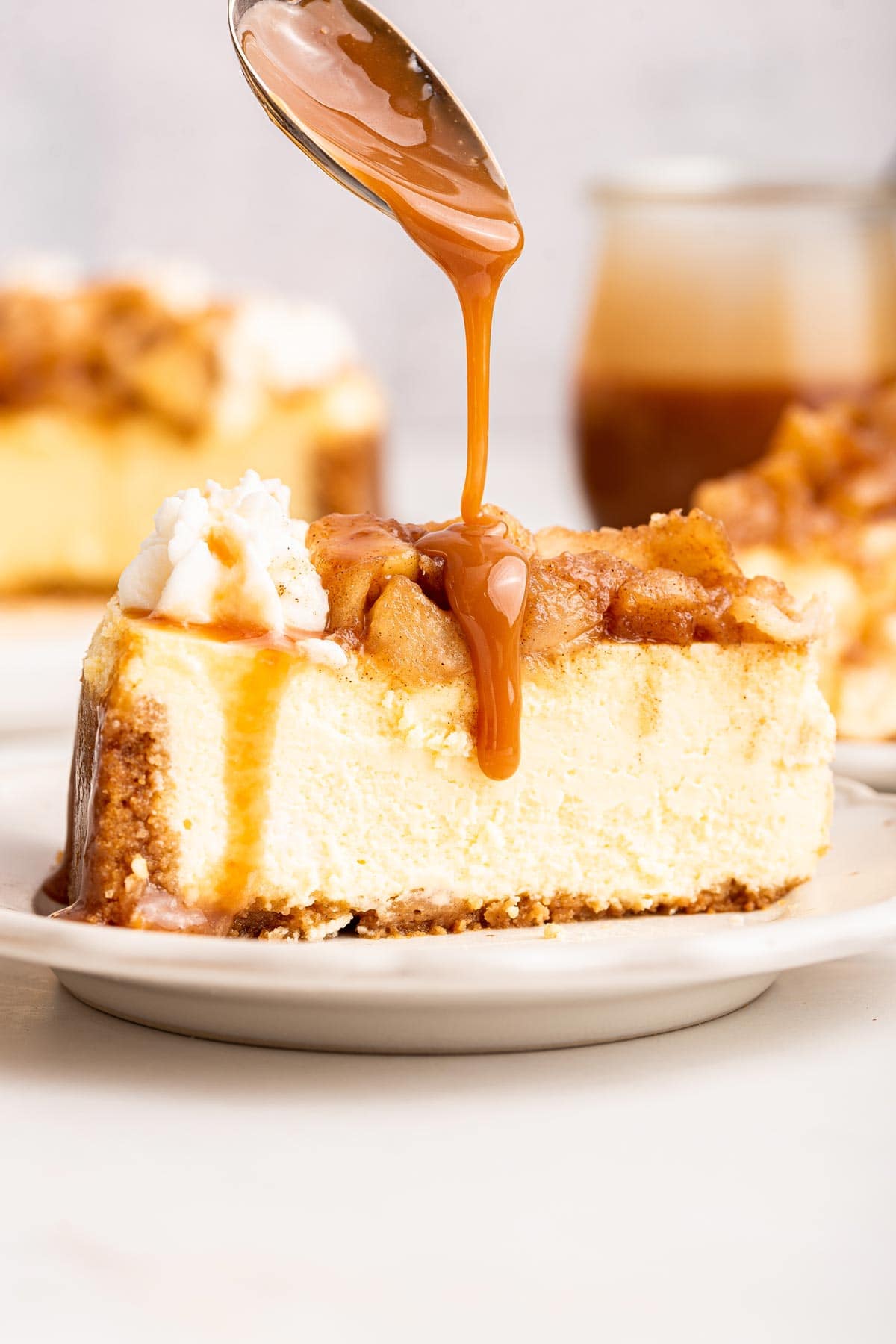 side view of a slice of cheesecake with a spoon drizzling caramel sauce on top.