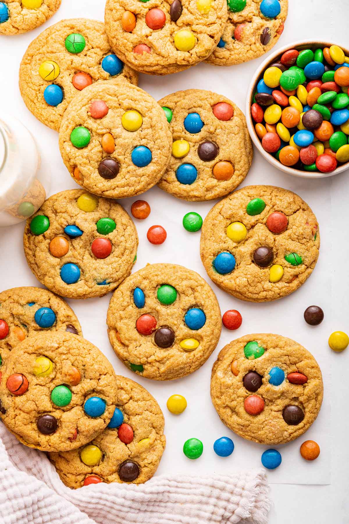 soft M&M cookies surrounded by additional m&ms and a glass of milk.
