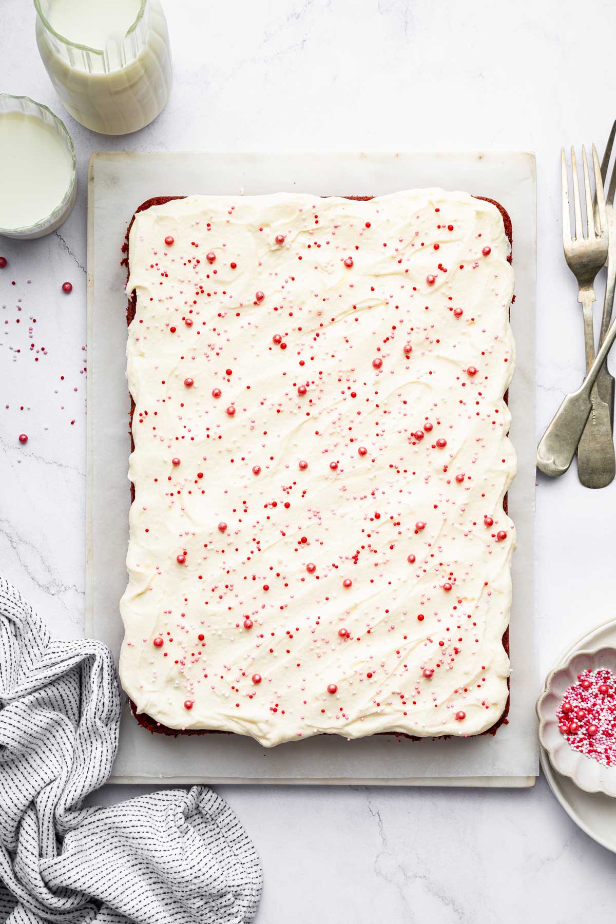 overhead view of a red velvet sheet cake covered with cream cheese frosting and red sprinkles.