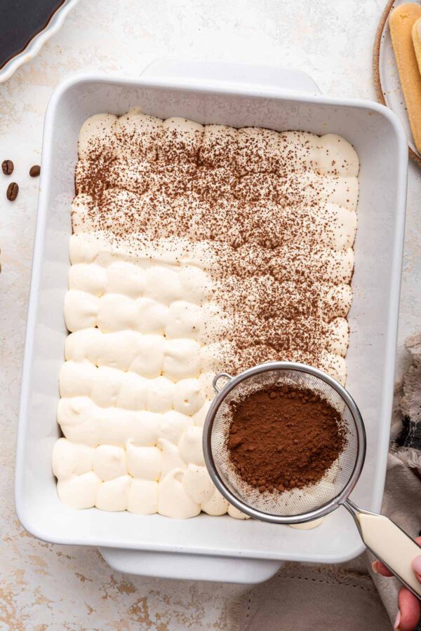 dusting homemade tiramisu with cocoa powder.