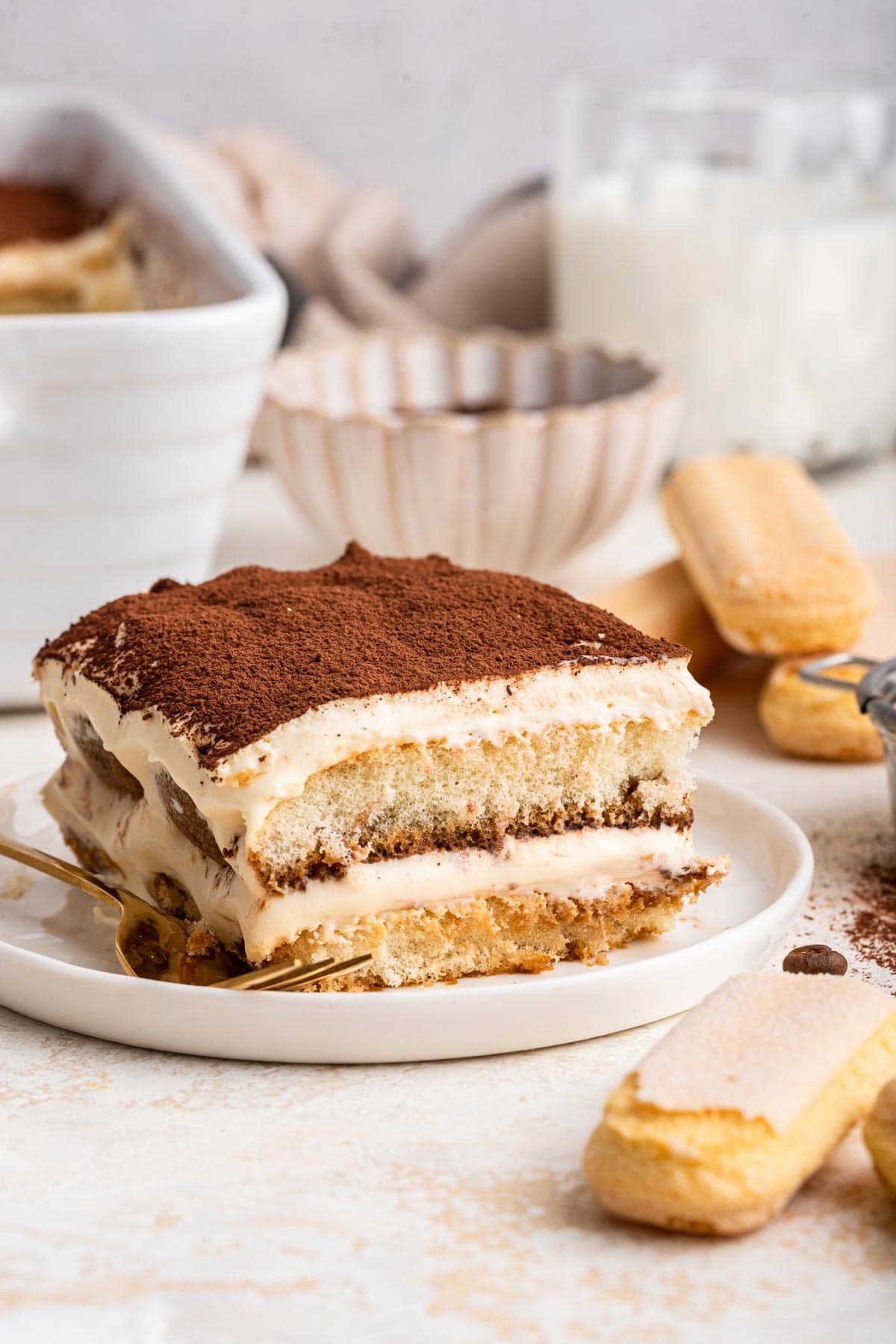 side view of a slice of tiramisu next to ladyfingers and coffee beans.
