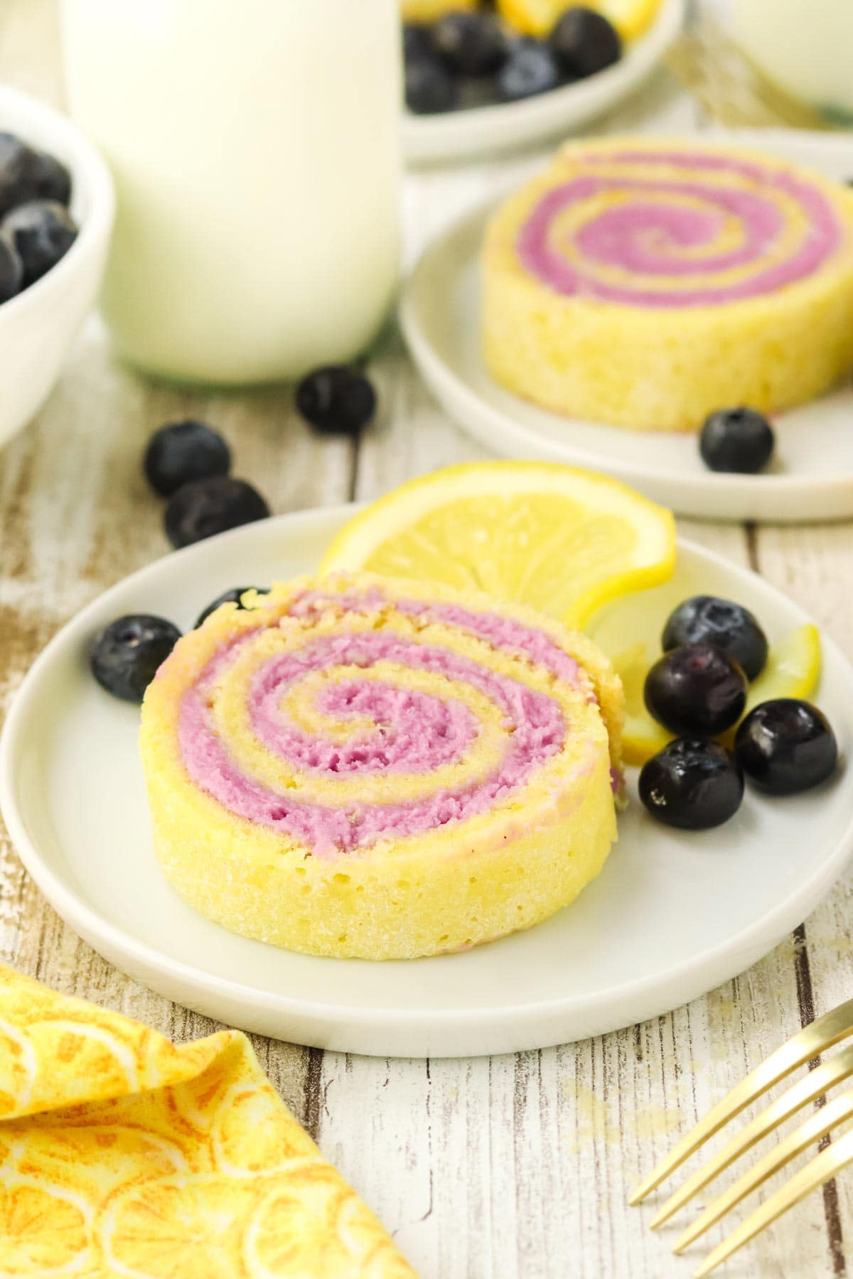 slice of spiral lemon blueberry cake roll on a plate next to blueberries and lemon slides.