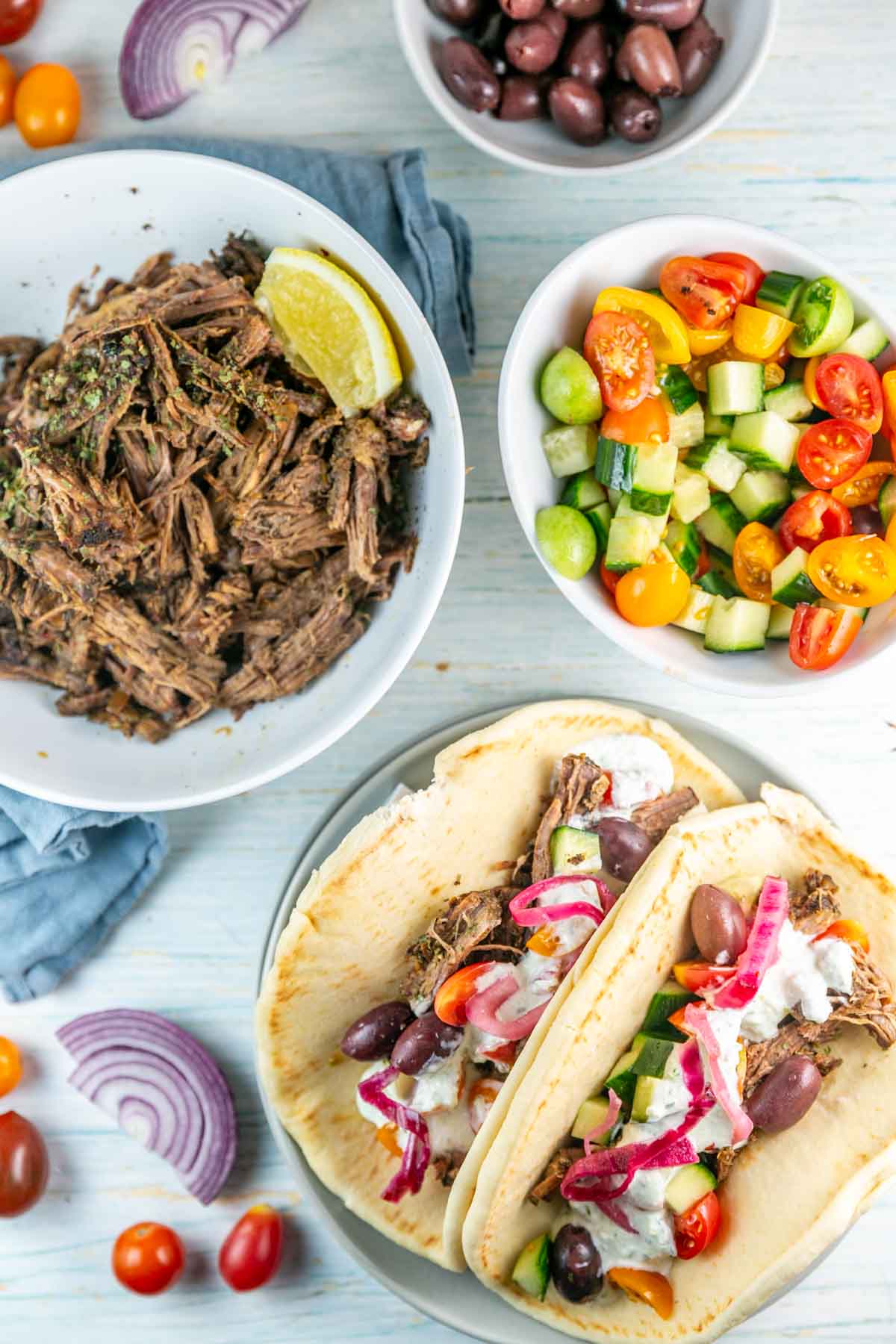 a large bowl of shredded beef next to tomato and cucumber salad and pitas to make gyros.