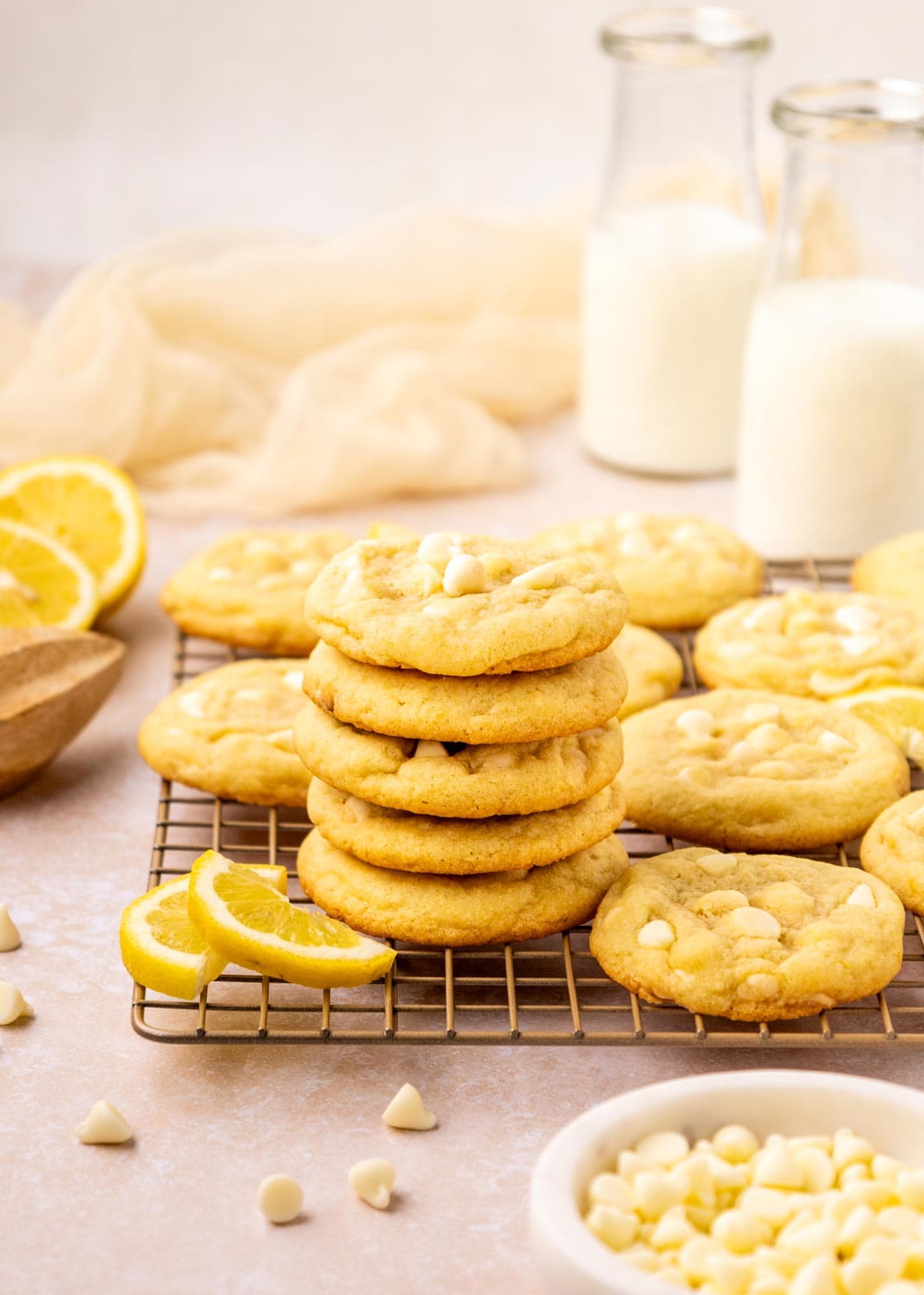 cookies stacked on a cooling rack surrounded by glasses of milk and white chocolate chips.