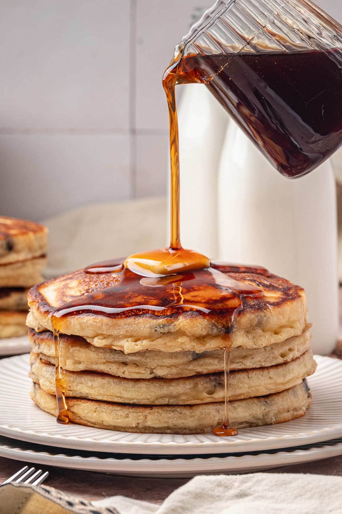 pouring maple syrup over a tall stack of chocolate chip pancakes.