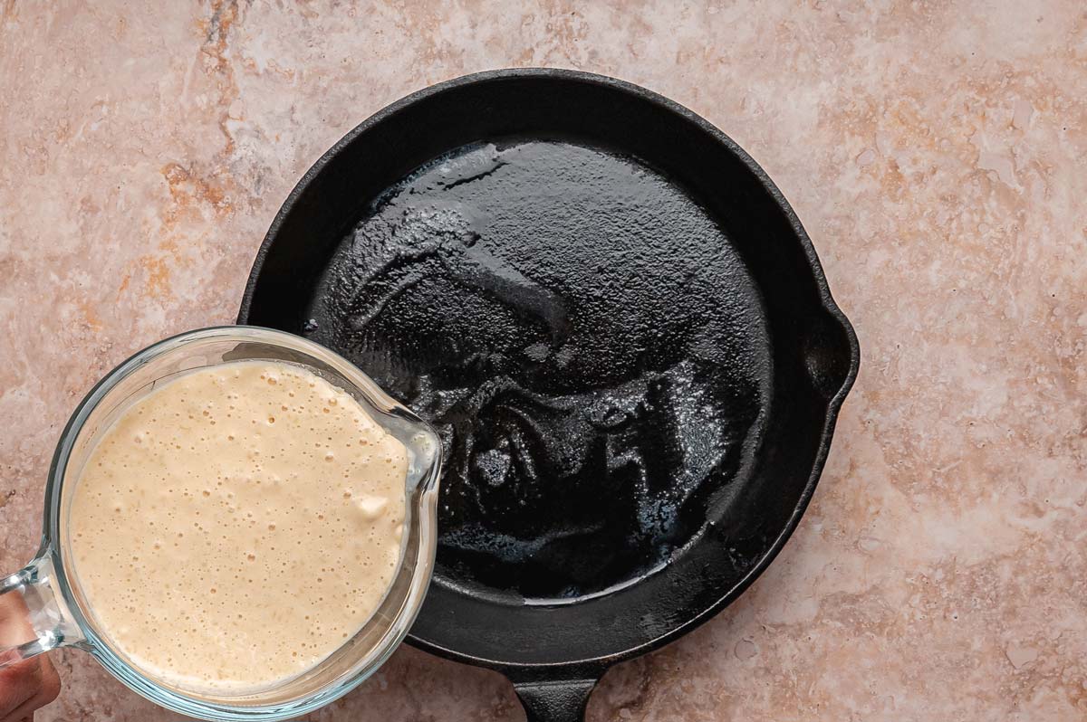pouring pancake batter into a cast iron skillet.